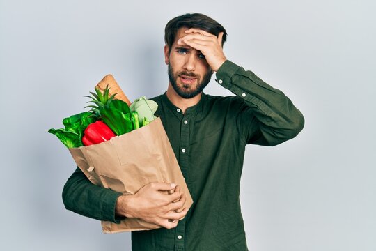 Young hispanic man holding paper bag with bread and groceries stressed and frustrated with hand on head, surprised and angry face
