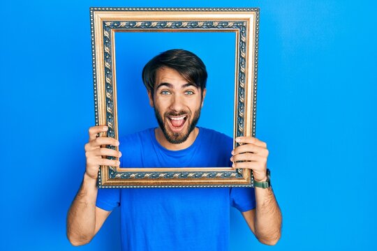 Young hispanic man holding empty frame smiling and laughing hard out loud because funny crazy joke.