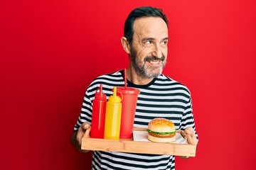 Middle age hispanic man eating a tasty classic burger and soda smiling looking to the side and staring away thinking.