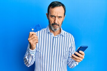 Middle age hispanic man using smartphone and holding credit card skeptic and nervous, frowning upset because of problem. negative person.