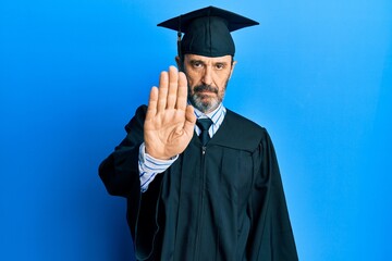 Middle age hispanic man wearing graduation cap and ceremony robe doing stop sing with palm of the hand. warning expression with negative and serious gesture on the face.