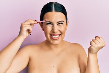Fototapeta premium Beautiful brunette woman brushing eyebrow with brush screaming proud, celebrating victory and success very excited with raised arm