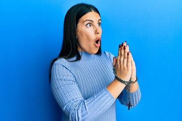 Beautiful brunette woman praying holding catholic rosary afraid and shocked with surprise and...