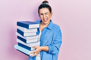 Young hispanic woman wearing glasses and holding books winking looking at the camera with sexy expression, cheerful and happy face.