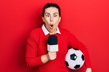 Young hispanic woman holding reporter microphone and soccer ball afraid and shocked with surprise...