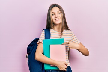 Young brunette girl holding student backpack and books smiling happy pointing with hand and finger