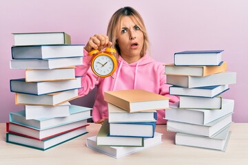 Young caucasian woman holding alarm clock sitting on the table with books in shock face, looking...