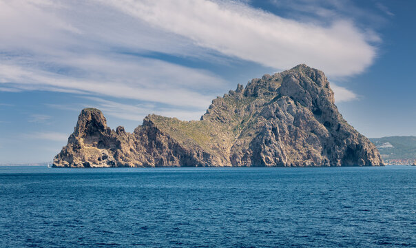 The small island of Vedra off the coast of Ibiza in the Mediterranean Sea. The rock rises steeply from the sea. On the left is a sailing boat. It's a sunny midday in summer with interesting clouds.