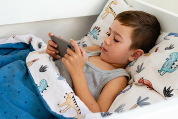 Little boy in bed  looking at the smartphone at morning. Teen boy lying on front in the bed and using smartphone. 5 years old child boy using smartphone at morning in bed