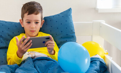 Little boy in bed  looking at the smartphone at morning. Teen boy lying on front in the bed and using smartphone. 5 years old child boy using smartphone at morning in bed