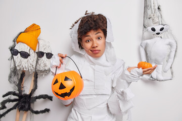 Indoor shot of hesitant clueless curly haired woman holds two halloween pumpkins dressed in ghost...
