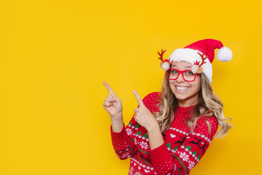 A Young Smiling Blonde Woman In A Red Christmas Sweater, Glasses With Deer Antlers And A Red Santa's Hat Points At Copy Space For Text Or Design With Her Fingers Isolated On A Color Yellow Background 