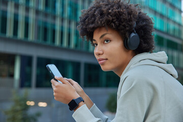 Sideways shot of thoughtful woman athlete with curly hair chooses song from playlist in smartphone looks away listens music in wireless headphones uses wearable smartwatch for tracking activity