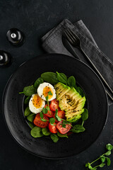 Avocado, cherry tomato, spinach and chicken egg, microgreens peas and black sesame seeds fresh salad in black bowl on black slate, stone or concrete background. Healthy food concept. Top view.