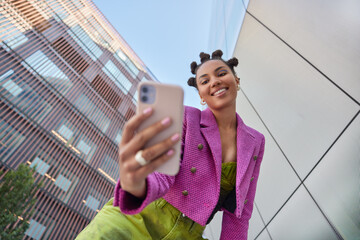 Happy young woman with hairstyle dressed in fashionable clothes makes video call or selfie via smartphone uses mobile app strolls at urban setting films vlog for social networks. Communication - Powered by Adobe