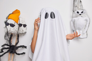 Confused mysterious female ghost has phone conversation keeps smartphone near ear poses against white studio background with spooky creatures around. Spooky holiday halloween celebration concept