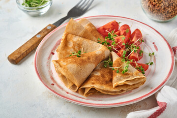 Stack of gluten free buckwheat flour crepes pancakes with cherry tomatoes and arugula microgreens on white plate, homemade healthy baking for breakfast. Mock up. Copy space.