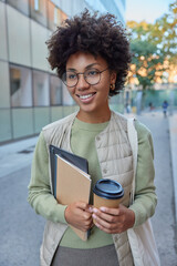 Vertical shot of attractive curly female model smiles pleasantly carries paper cup of coffee digital tablet and notebook wears optical glasses for vision correction strolls outside during daytime