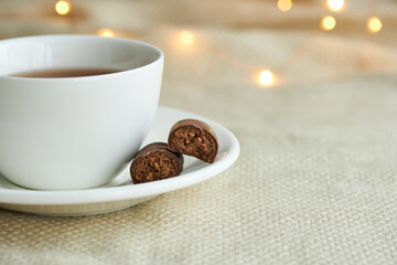 White cup of tea with chocolate sweets on a beige plaid. Christmas lights and copy space.