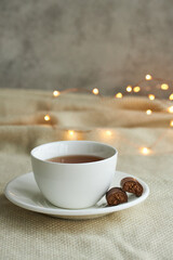 White cup of tea with chocolate sweets on a beige plaid. Christmas lights and copy space.