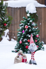 Snowy handmade dwarf in red white clothes with bag of gifts in decorated Christmas tree background in winter time in yard, winter holiday concept