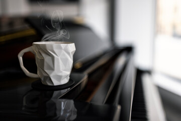 A white edgy porcelain coffee cup with steam rising from the heat on a black glossy grand piano