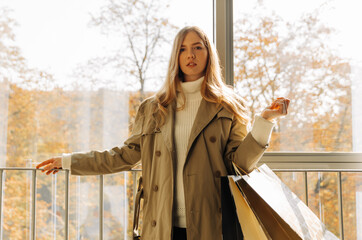 woman in a beige raincoat walks through the mall with shopping bags in her hands, in the autumn...