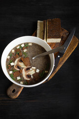 Vegan mushroom cream soup with toasted bread, dill and smoked tofu. A сut piece of smoked tofu and a knife on a wooden cuttong board. Dark wooden background, top view.