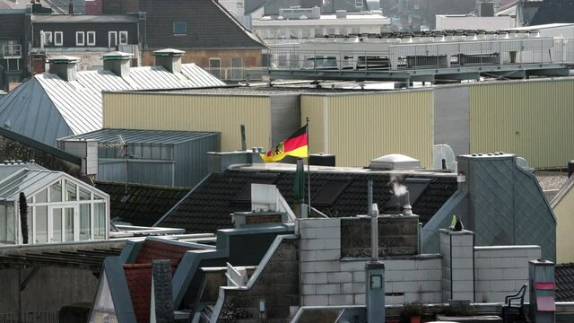 View Of A Roof In Aachen On Which A German Flag Is Flying In A Light Wind. Smoke Comes Out Of Small Chimneys. District Of North Rhine-Westphalia In Germany