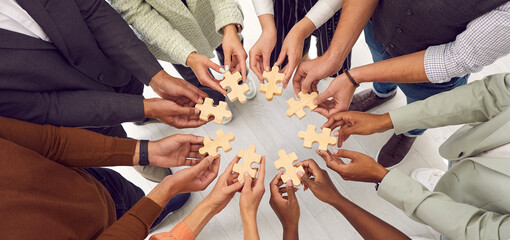 Banner background with multiethnic group of young people standing together and joining pieces of jigsaw puzzle as metaphor for business team and teamwork. Cropped shot of hands holding jigsaw parts