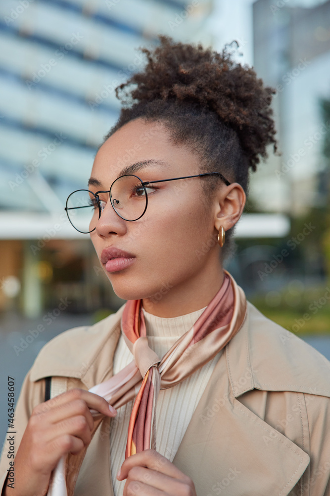 Wall mural Vertical shot of beautiful curly haired millennial gir wears round spectacles for vision correction coat and tied kerchief around neck looks thoughtfully forward while strolling outside during daytime