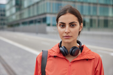 Photo of serious female sports trainer looks directly at camera strolls on road alone against urban background wears jacket headphones around neck leads healthy lifestyle exercises every day