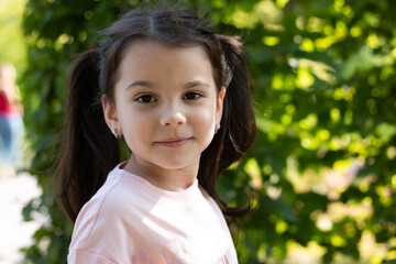 Little cheerful girl with ponytails smiling looks ahead against a background of green blurred foliage