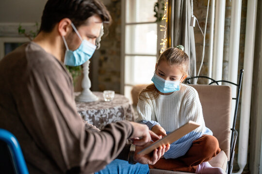 Father with disability in wheelchair using tablet at home with child while wearing masks.