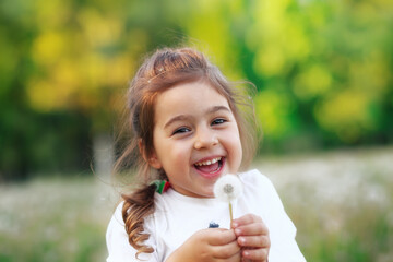 Cute little girl with dandelion flower is smiling in spring park. Happy cute kid having fun outdoors at sunset.