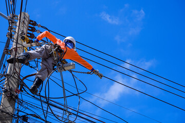 Low angle view of electrician with safety equipment and various work tools is installing cable lines and electrical system on electric power pole against blue sky background - obrazy, fototapety, plakaty