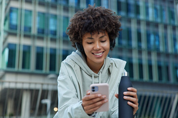 Positive sporty woman drinks water after exercising uses mobile phone scrolls social networks smiles happily dressed in hoodie and anorak poses against modern city building enjoys messaging. © WHstudio Leushin N