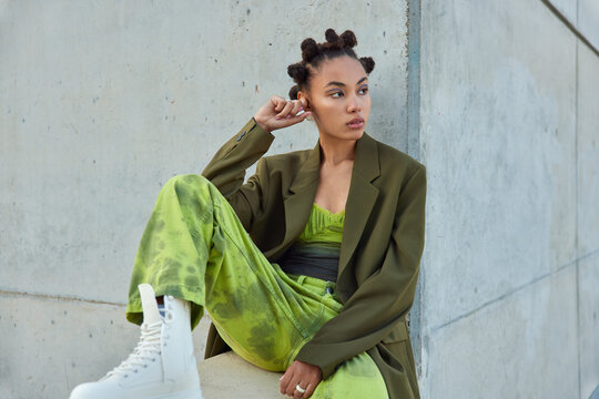 Horizontal shot of beautiful teenage girl with trendy hairstyle dressed in green clothes looks away poses against urban grey wall considers something. People youth lifestyle and fashion concept
