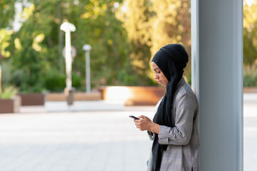 Side view of a muslim adult woman leaning on a support beam column looking down while using her phone