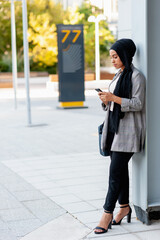 Side view of an adult woman leaning on a support beam column looking down while using her phone