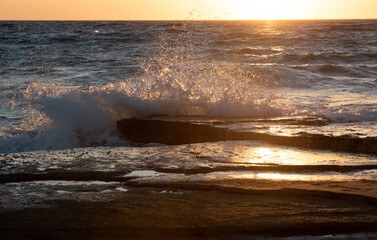 Naklejka premium Stormy windy sea waves splashing on a rocky seashore at sunset