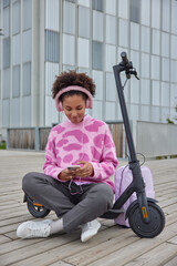 Relaxed teenage girl sits crossed legs near electric scooter rests after driving listens music via earphones and smartphone enjoys leisure time in city surfs social networks. People lifestyle hobby
