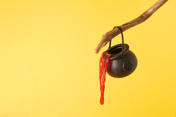 black small witch cauldron with artificial decorative blood on a branch, yellow background,...