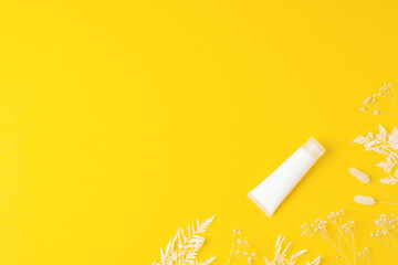 White tube of cosmetic cream with flowers on yellow background. Flat lay, copy space