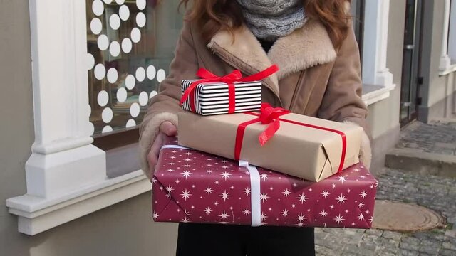 Teengirl in grey hat, scarf and brown jacket holding a set of christmas gifts near shops window. Happy young woman preparing holidays presents. Concept of shopping, giving and receiving gifts.