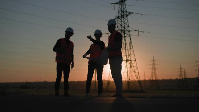 professional men and woman with project in their hands are discussing structure of lines of an electric tower on background of sunset, silhouette