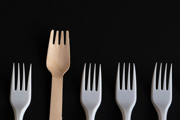 Wooden disposable fork made from birch tree rises above plastic ones lying on black background. Focus on wooden fork. Copy space for your text. Biodegradable cutlery. Environmental protection theme.