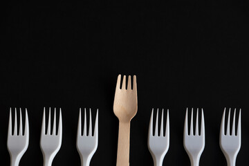 Wooden disposable fork made from birch tree rises above plastic ones lying on black background. Focus on wooden fork. Copy space for your text. Biodegradable cutlery. Environmental protection theme.