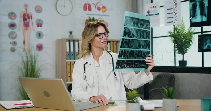 Medicine concept where beautiful confident smart qualified blond female doctor in glasses filling medical card on laptop using results of x-ray scan ,front view