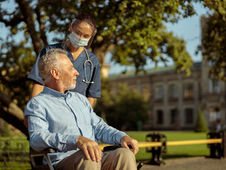 Caring young nurse wearing face shield and mask talking to mature man, pushing patient in wheelchair